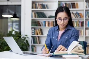 Girl with Glasses Studying in Library with Books and Laptop