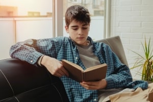 Young Boy Reading Book Sitting on Sofa at Home