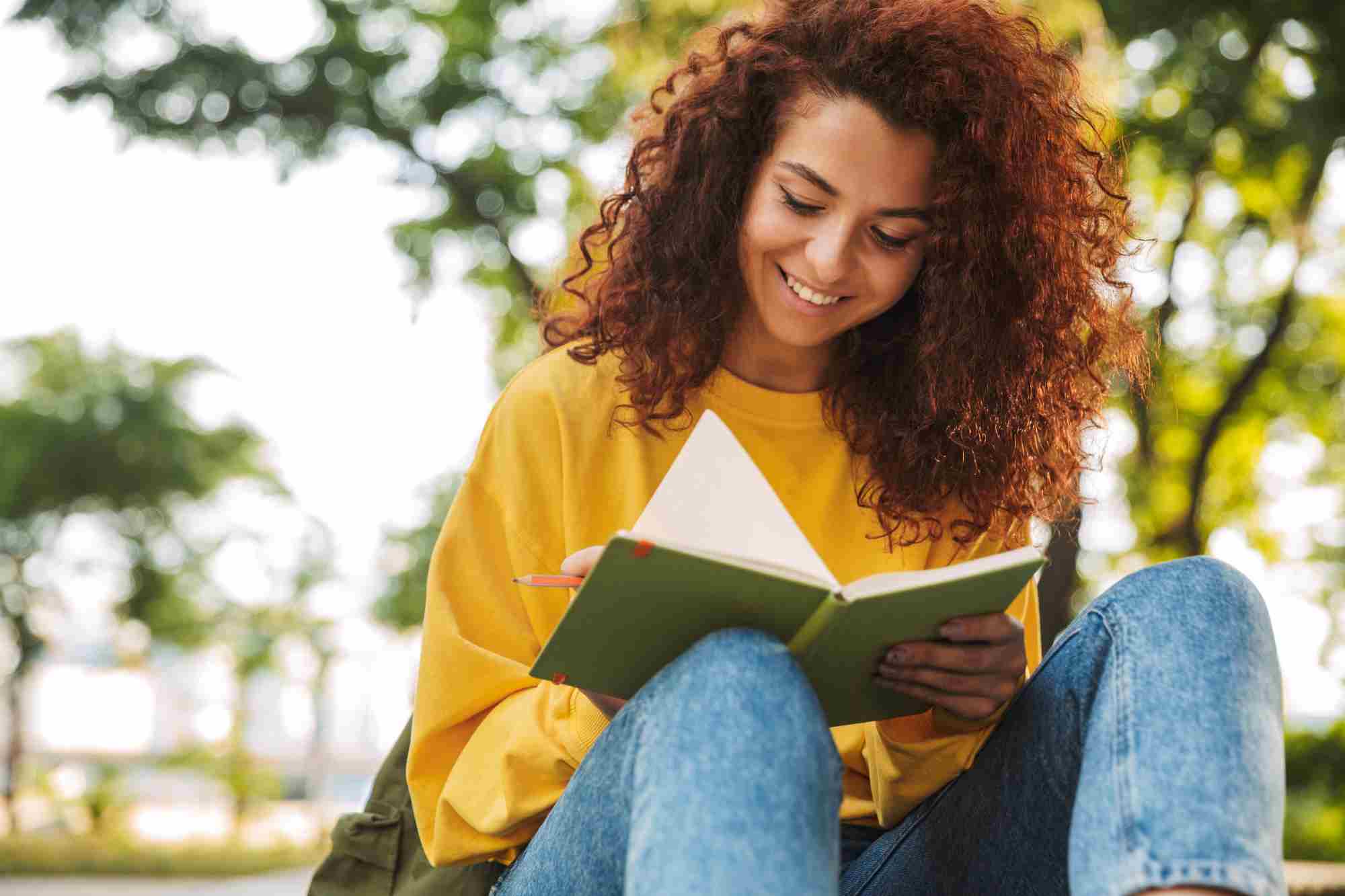 Curly Girl Sitting Outdoors Writing Notes in Notebook
